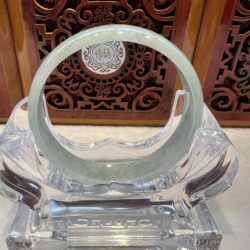 Clear圆形 glass sculpture resting on a faceted base, set against an ornate carved wooden panel with scrollwork in the background.