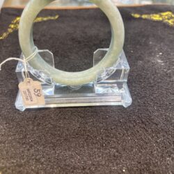 Light green jade bangle bracelet on a clear acrylic display stand in a jewelry shop, tag attached at the side.