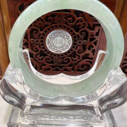 Pale green jade circular ring resting against an ornate dark wood panel with intricate scrollwork and a central silver medallion with Chinese characters.